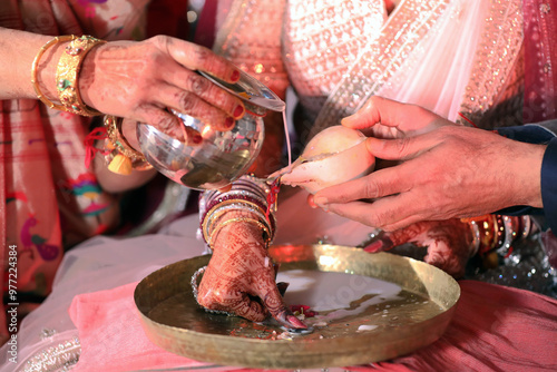 A traditional Indian wedding ritual, with the couple's hands adorned in henna and jewelry, pouring sacred water into a brass dish as part of a ceremonial offering. photo