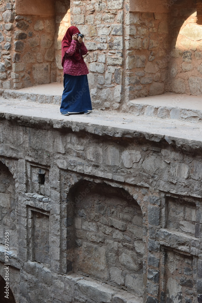 A girl in a maroon hijab stands firm in the stepwell of the famous Ugrasen ki Baoli in Delhi, capturing the historic architecture with her camera against a backdrop of ancient stone steps