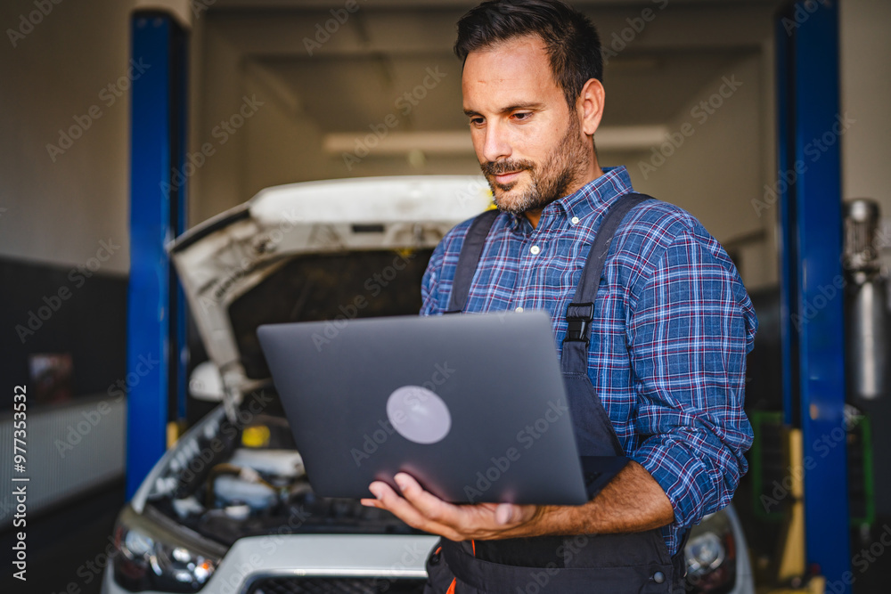 Adult man car mechanic work on laptop in garage with car in background Adult man car mechanic work on laptop in garage with car in background