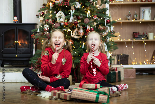Cute girls sitting next to a Christmas tree on the floor and eating gift sweets