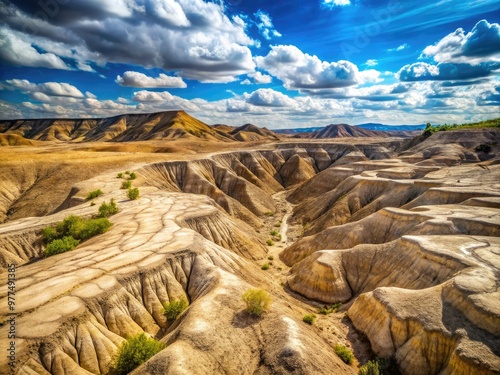 Rugged, barren hillside exhibits severe erosion, with deep gullies and ravines etched into the dry, cracked earth, against a stark blue sky with few clouds. photo