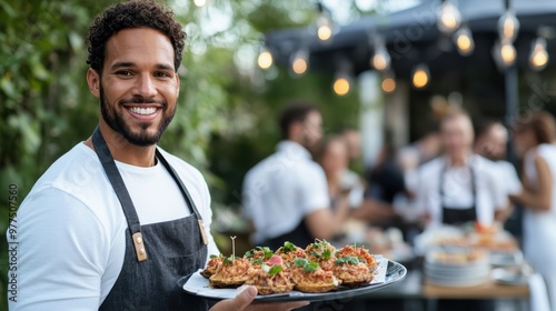 A smiling chef in a white t-shirt and apron holds a meticulously arranged platter at an outdoor event, conveying joy and satisfaction in a vibrant social setting.