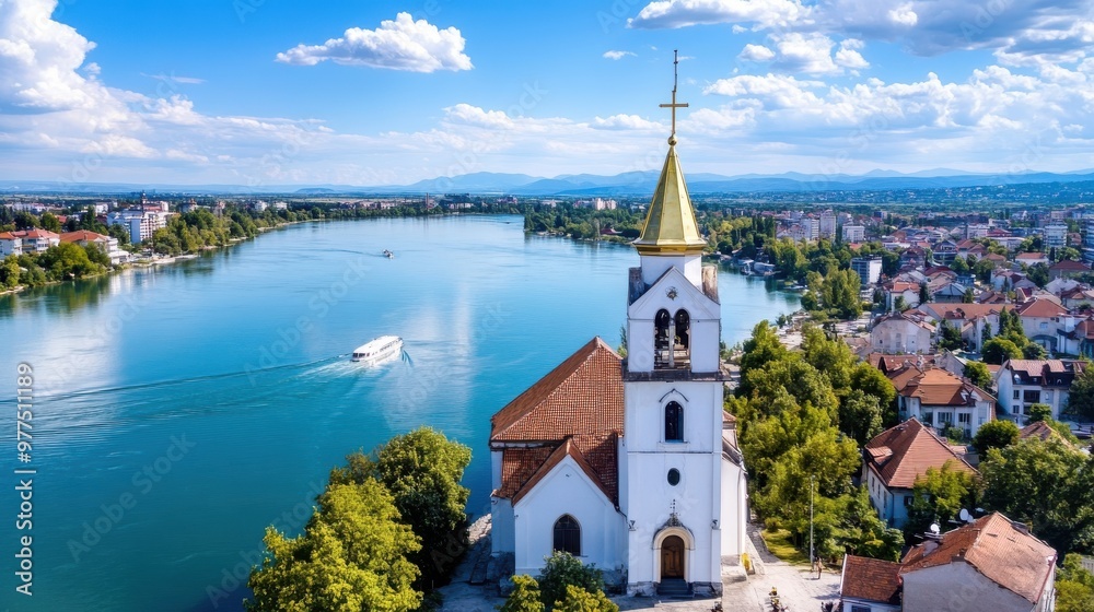 Fototapeta premium A picturesque riverside church with a golden spire overlooking a serene, wide river where a small boat sails by; framed by a clear, blue sky and historical architecture.