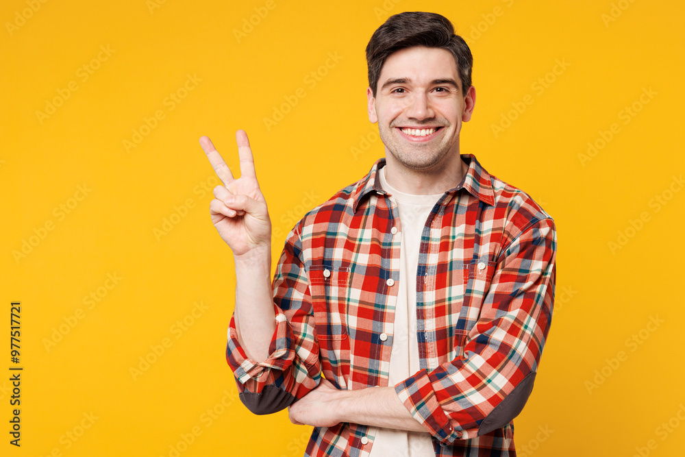Young smiling happy cheerful joyful man he wears red checkered shirt casual clothes showing victory sign looking camera isolated on plain yellow orange background studio portrait. Lifestyle concept. Young smiling happy cheerful joyful man he wears red checkered shirt casual clothes showing victory sign looking camera isolated on plain yellow orange background studio portrait. Lifestyle concept.