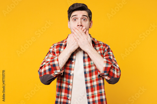 Young shocked surprised astonished sad man he wears red checkered shirt casual clothes cover mouth with hand look camera isolated on plain yellow orange background studio portrait. Lifestyle concept.