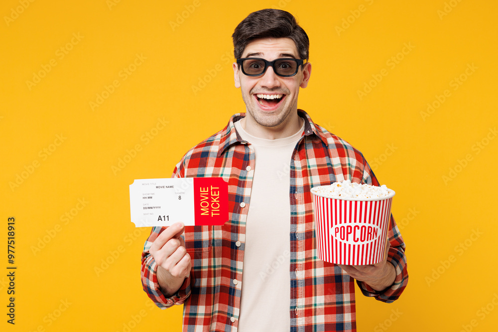 Young surprised happy man he wearing red checkered shirt casual clothes 3d glasses watch movie film hold ticket bucket of popcorn in cinema isolated on plain yellow orange background studio portrait. Young surprised happy man he wearing red checkered shirt casual clothes 3d glasses watch movie film hold ticket bucket of popcorn in cinema isolated on plain yellow orange background studio portrait.