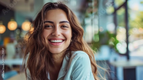 A smiling woman with curly hair is pictured in a light green shirt at a café. Background includes green plants and tables. No wind evident to suggest hair movement.