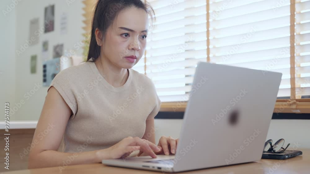 A woman is typing on a laptop in front of a window. She is wearing a tan shirt and has her hair in a ponytail A woman is typing on a laptop in front of a window. She is wearing a tan shirt and has her hair in a ponytail