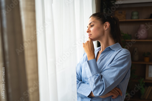 Fotografie Young woman living alone in apartment looking out the window spying on new neighbors moving into house or apartment across the street from hers