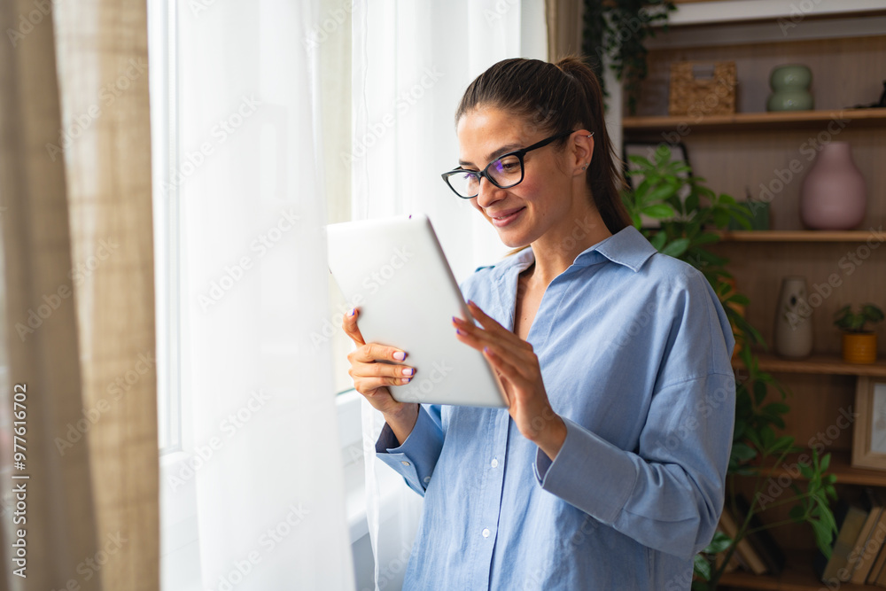Smart caucasian woman in business casual wear holding digital tablet standing near windows at home. Young freelance independent expert female worker using technology for distant work at home office. Smart caucasian woman in business casual wear holding digital tablet standing near windows at home. Young freelance independent expert female worker using technology for distant work at home office.