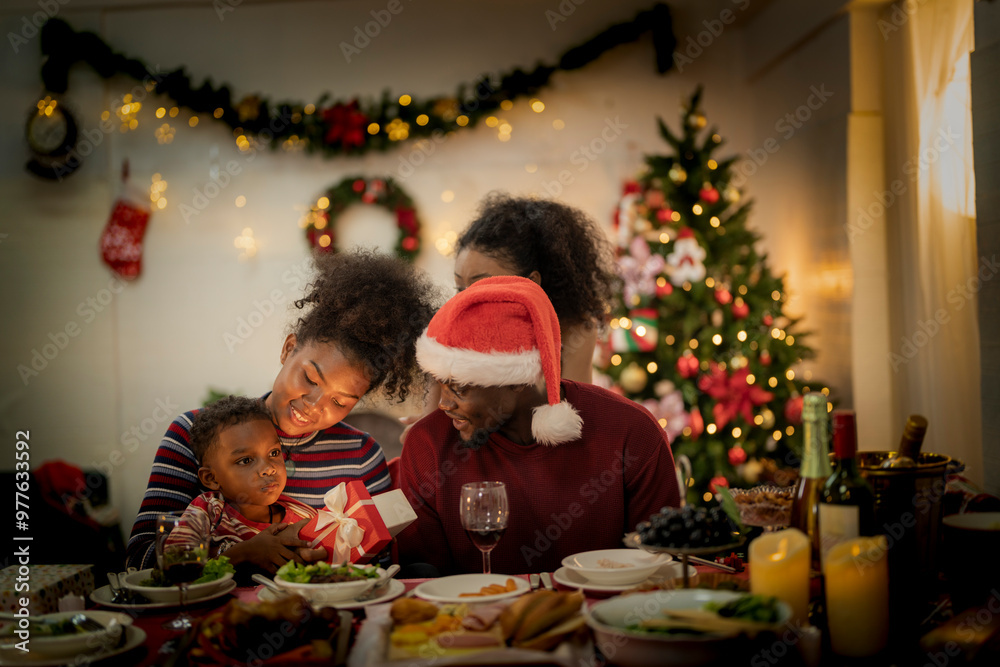 A joyful family gathers around the dinner table celebrating Christmas. The father, dressed in a Santa hat, gives a gift to the child, while everyone smiles with warmth and festive decorations. A joyful family gathers around the dinner table celebrating Christmas. The father, dressed in a Santa hat, gives a gift to the child, while everyone smiles with warmth and festive decorations.