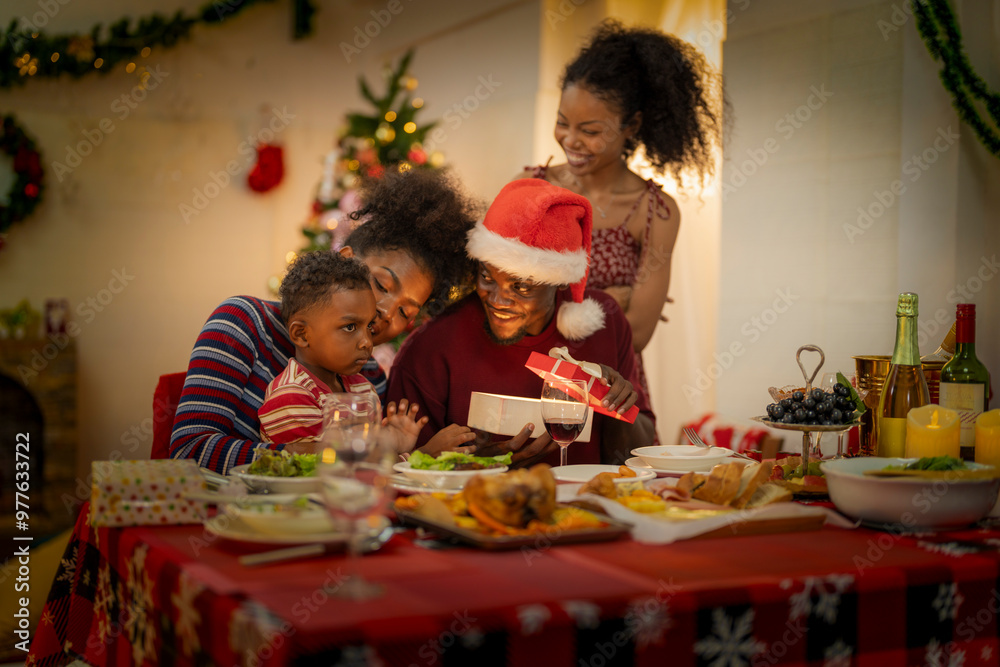 A joyful family gathers around the dinner table celebrating Christmas. The father, dressed in a Santa hat, gives a gift to the child, while everyone smiles with warmth and festive decorations. A joyful family gathers around the dinner table celebrating Christmas. The father, dressed in a Santa hat, gives a gift to the child, while everyone smiles with warmth and festive decorations.