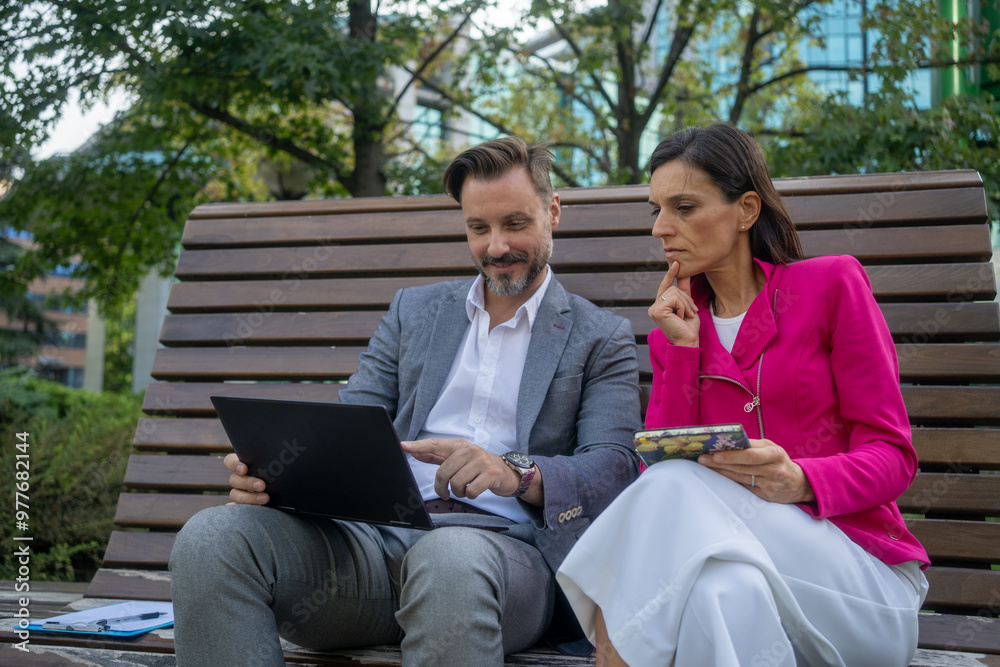 Team of diverse partners sitting at bench mature Latin business man and European business woman discussing project on laptop at park. Two colleagues of professional business people working together. Team of diverse partners sitting at bench mature Latin business man and European business woman discussing project on laptop at park. Two colleagues of professional business people working together.