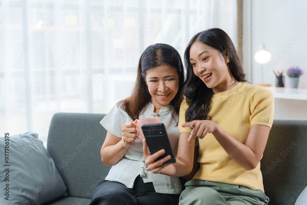 Happy asian mother and adult daughter bonding on the couch at home, using a mobile phone to browse social media together Happy asian mother and adult daughter bonding on the couch at home, using a mobile phone to browse social media together