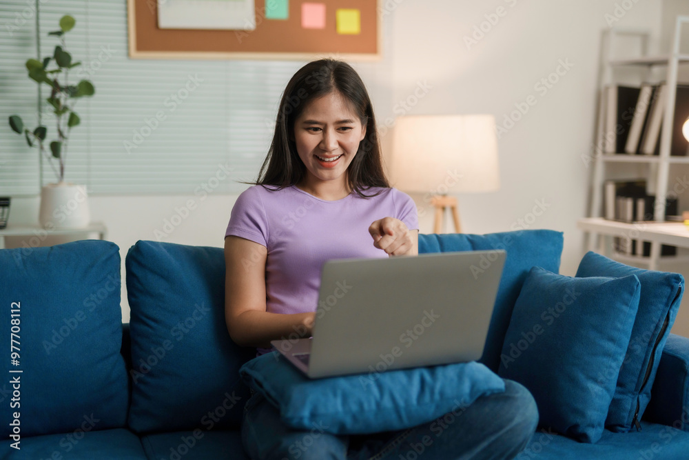 Young woman sitting on a blue couch, pointing and smiling while using a laptop for a video call Young woman sitting on a blue couch, pointing and smiling while using a laptop for a video call