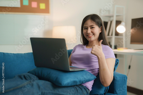 Young asian woman relaxes on her sofa at home, happily using a laptop and giving a thumbs up. She looks content, fully immersed in technology and the internet, working and browsing online