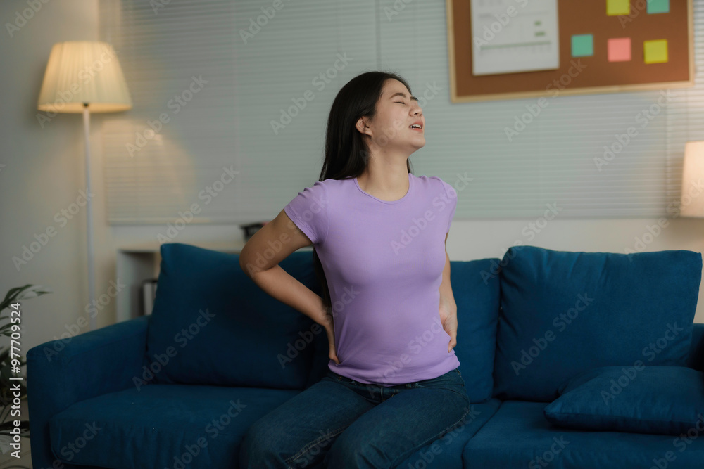 Young woman grimacing in pain as she sits on her sofa at home, holding her back due to back pain Young woman grimacing in pain as she sits on her sofa at home, holding her back due to back pain