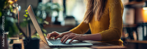 A woman wearing a yellow sweater is focused on typing on her laptop in a cozy indoor setting with natural light. photo