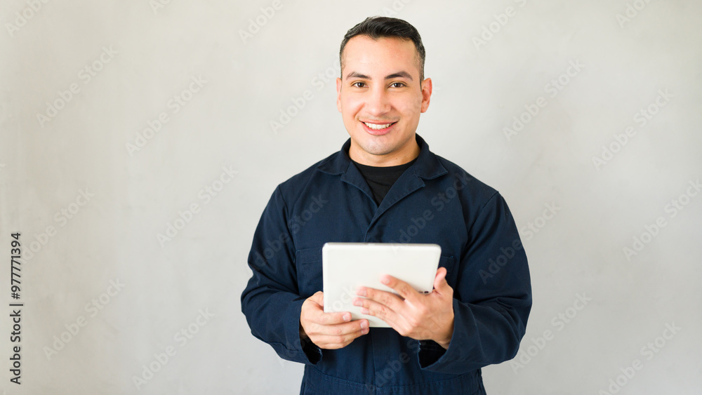 Smiling mechanic in blue overalls holds a tablet in a bright studio, radiating confidence and expertise in automotive repair Smiling mechanic in blue overalls holds a tablet in a bright studio, radiating confidence and expertise in automotive repair