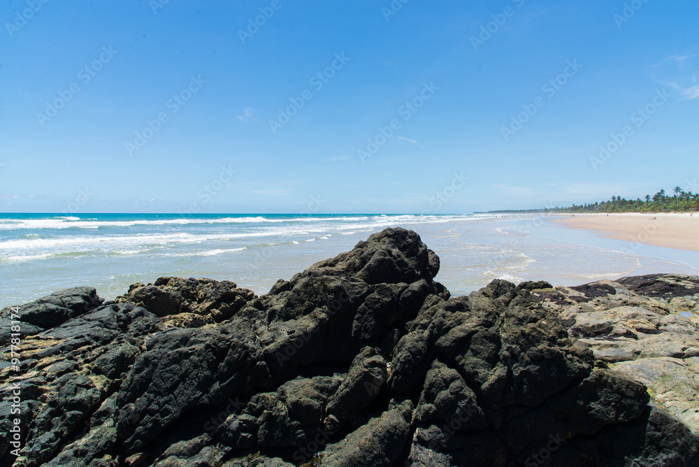 View of a beautiful beach with blue waters and dark rocks, against a clear sky. View of a beautiful beach with blue waters and dark rocks, against a clear sky.