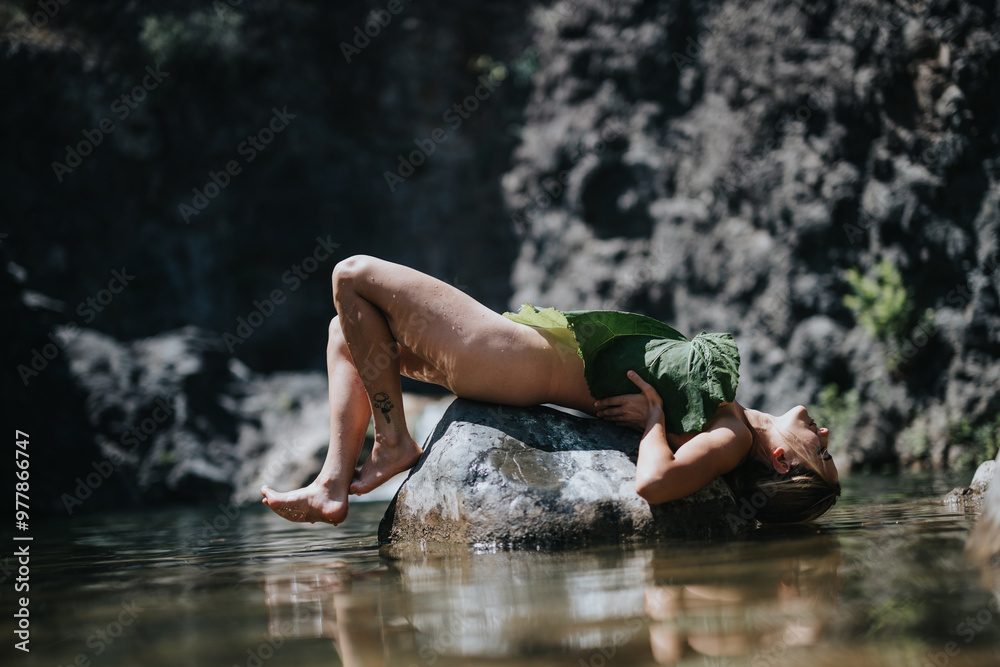 Fototapeta premium A woman lies on a rock in a serene outdoor setting, covered with a large leaf. The image conveys relaxation and connection with nature.