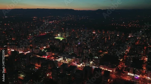 Birds-eye view shows a city slowly being enveloped in darkness as the sun sets. Street lights and lights from buildings illuminate the urban landscape