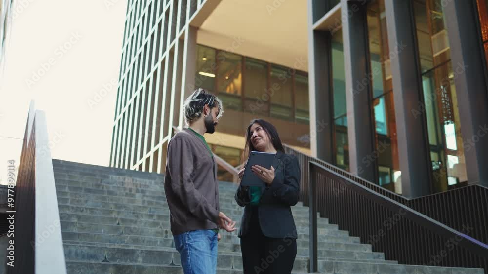 Businesswoman explaining contract details on a digital tablet to her business partner while standing on stairs against a large office building in the background. Businessmen discussing details Businesswoman explaining contract details on a digital tablet to her business partner while standing on stairs against a large office building in the background. Businessmen discussing details