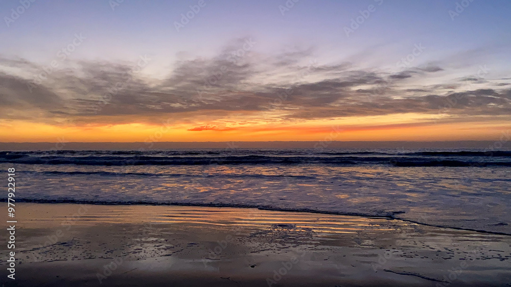 Fototapeta premium Orange and pink skies as sun goes down overlooking sandy beach while ocean tides create waves