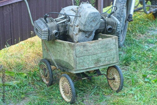 one rusty heavy iron retro dirty mechanical power gray engine repaired from an old two-cylinder classic retro motorcycle lies in a small four wheeled wooden green cart during the day on the street 