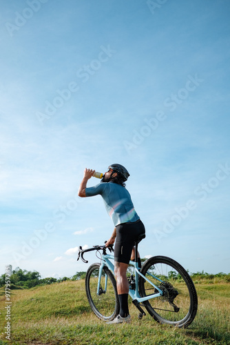 A man drinking from his bottle with his grave bike in the open fields.