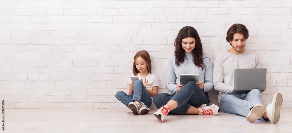 Fototapeta premium Family Gadgets. Happy parents and their little daughter holding different electronic devices in hands while sitting on floor near white brick wall