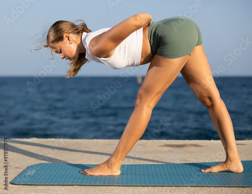 On windy day young slender woman does yoga near sea on embankment doing asanas, performs physical exercises. During sunset, girl performs Parshvottanasana photo