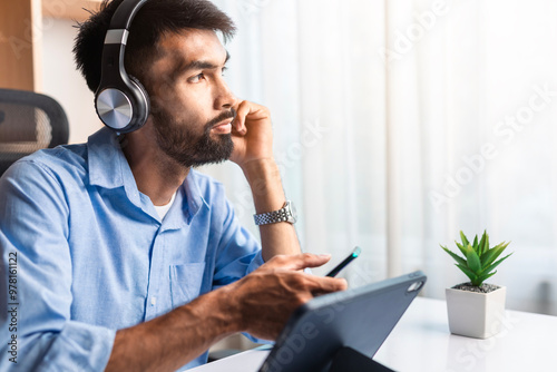 Serious young businessman with beard listening to music on headphone, looking outside while working in office.