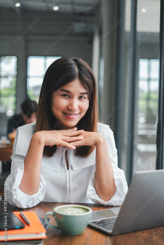 A young professional woman smiling at the camera while working on her laptop in a modern cafe setting. A young professional woman smiling at the camera while working on her laptop in a modern cafe setting.