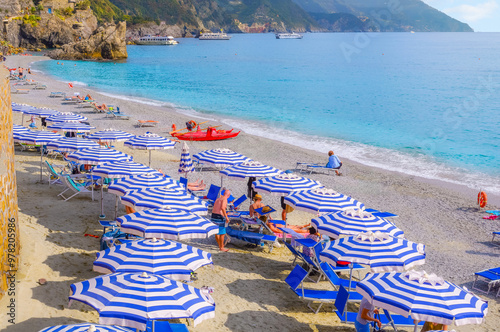 Fototapeta Naklejka Na Ścianę i Meble -  The sandy beach at Monterosso Al Mare, Cinque Terre Italy, with blue and white striped umbrellas, sunbathers, the Ligurian sea and boats along the rocky coast.