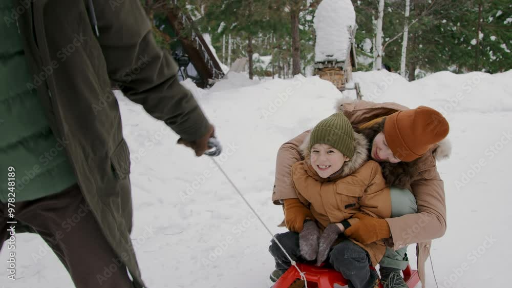 Medium full tracking shot of joyous young mother and son riding in sled together in snow in winter forest during holidays, hugging, cheering and laughing, and unrecognizable father pulling them Medium full tracking shot of joyous young mother and son riding in sled together in snow in winter forest during holidays, hugging, cheering and laughing, and unrecognizable father pulling them