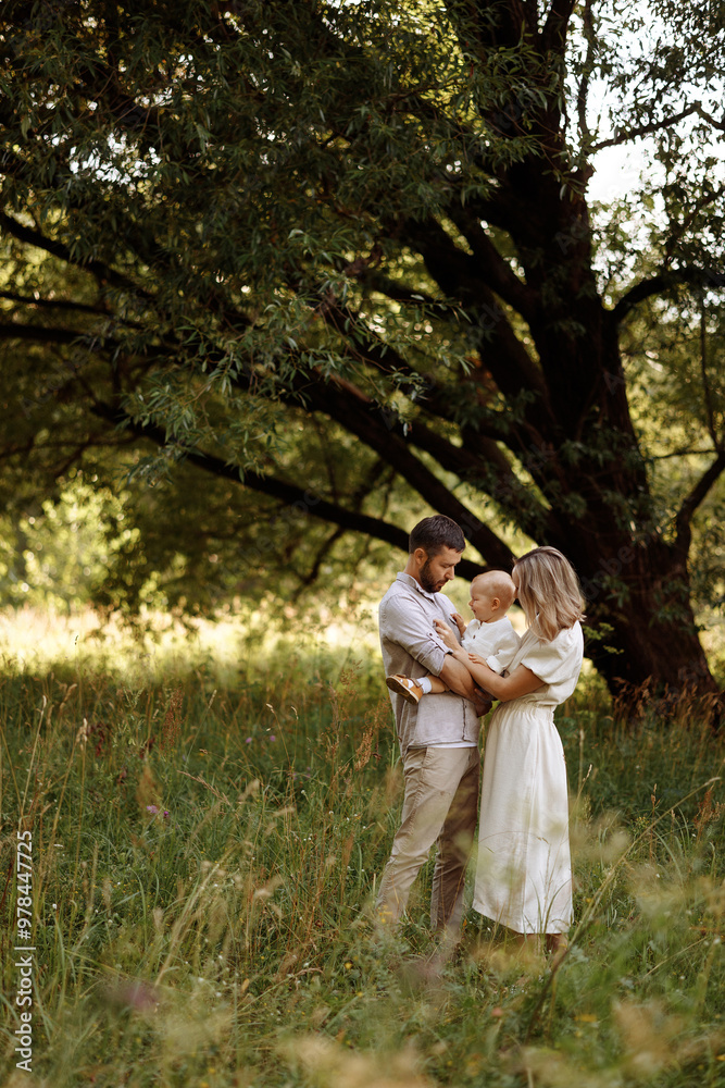 A family of three, a man and a woman with a child in their arms, stand in a park in the summer. The concept of a happy family with a small child, care and love.