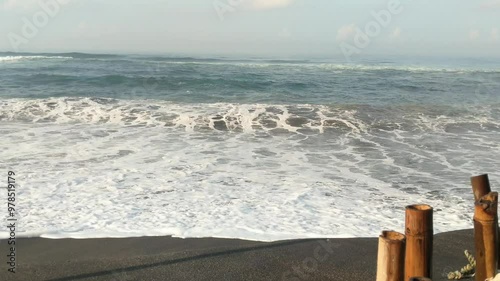 A vast ocean with large, rolling waves. The waves are crashing onto a sandy beach, creating white foam. The sky is blue with white clouds. In the distance, there are mountains or hills covered in fog. photo