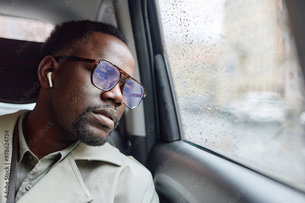Man wearing glasses and earbuds, sitting in a car on a rainy day, staring out window with a pensive expression, wearing beige jacket Man wearing glasses and earbuds, sitting in a car on a rainy day, staring out window with a pensive expression, wearing beige jacket