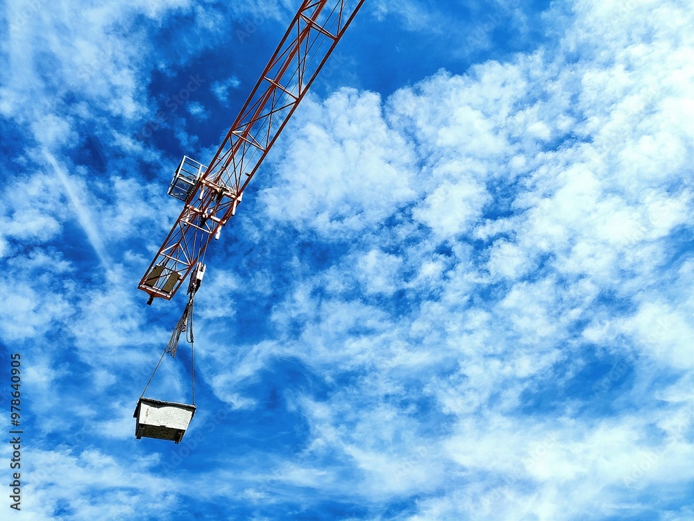 A towering construction crane stands against a backdrop of blue sky and scattered clouds. Its orange steel framework and intricate design suggest heavy lifting capacity, with reinforced beams and safe A towering construction crane stands against a backdrop of blue sky and scattered clouds. Its orange steel framework and intricate design suggest heavy lifting capacity, with reinforced beams and safe