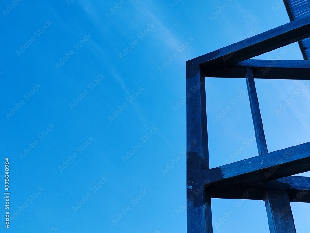 A steel framework stands against a clear blue sky, with sharp lines and geometric shapes, creating a modern and minimalistic architectural composition. The dark metal contrasts starkly with the bright A steel framework stands against a clear blue sky, with sharp lines and geometric shapes, creating a modern and minimalistic architectural composition. The dark metal contrasts starkly with the bright
