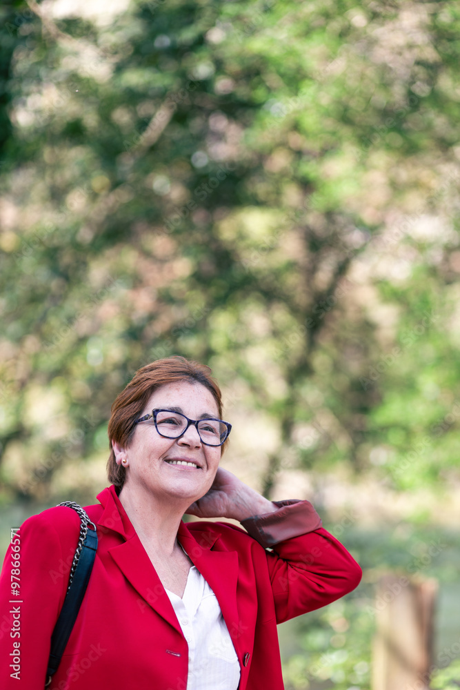A mature woman with glasses smiles warmly while wearing a red blazer. The background is blurred, with lush green foliage and sunlight filtering through. A mature woman with glasses smiles warmly while wearing a red blazer. The background is blurred, with lush green foliage and sunlight filtering through.