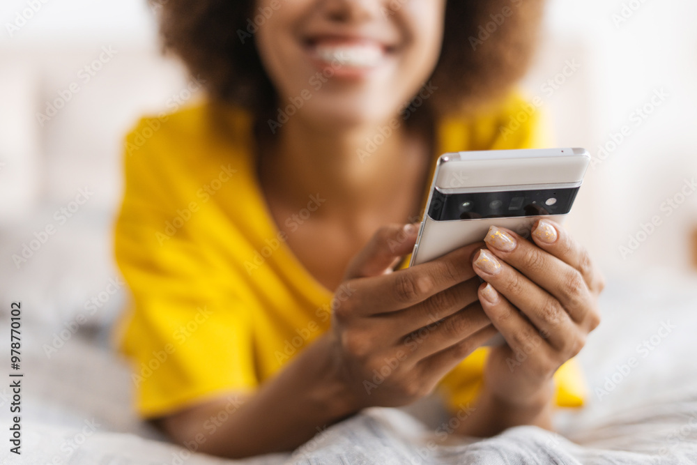Close up of smiling African American woman holding mobile phone lying on bed Close up of smiling African American woman holding mobile phone lying on bed