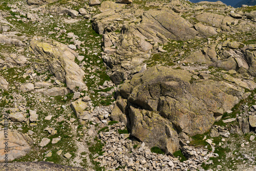 A detailed view of a rocky terrain featuring large boulders and scattered stones, interspersed with patches of green vegetation. The rugged landscape photo