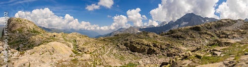 A panoramic view of a rugged mountain landscape featuring rocky terrain and patches of greenery under a clear blue sky with scattered clouds