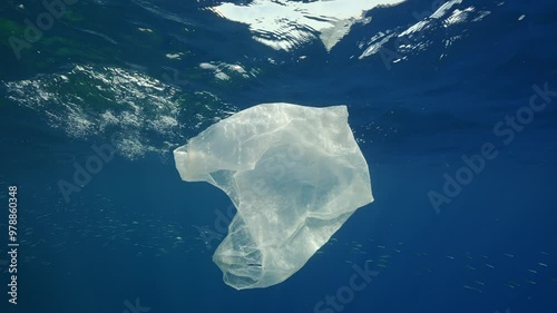 Discarded disposable white plastic bag slowly swims underwater reflected on surface, school of fish swims on background, Mediterranean sea in bright sun beams, Slow motion 