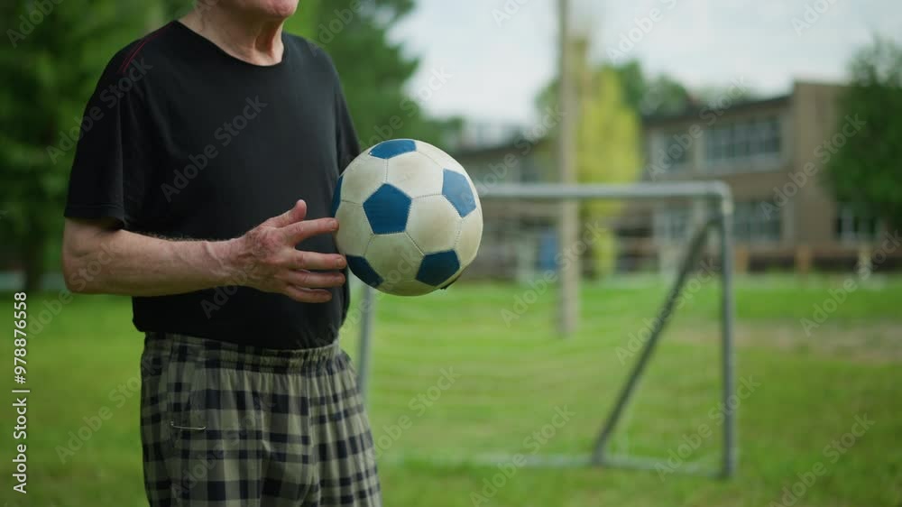 An elderly man rolls a soccer ball back and forth in his hand with focused intent, in the blurred background, a goalpost and a building can be seen An elderly man rolls a soccer ball back and forth in his hand with focused intent, in the blurred background, a goalpost and a building can be seen
