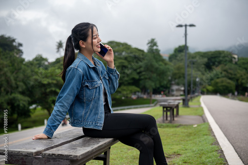 A positive Asian woman in a denim jacket is talking on the phone with someone on a bench in a park.