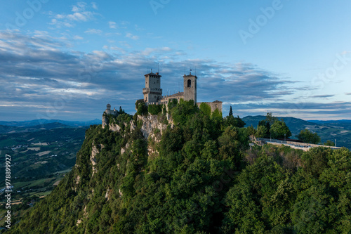 Aerial view of a historic medieval castle and tower on a hilltop surrounded by lush green forest at sunset, Borgo Maggiore, San Marino.