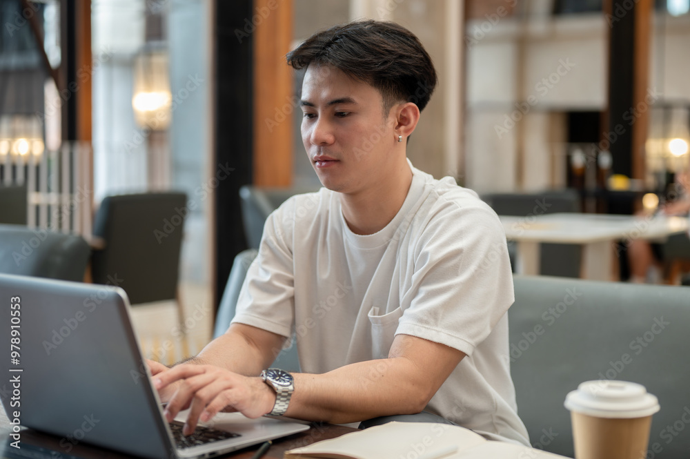 © bongkarn - A handsome, focused Asian man working remotely from a coffee shop, working on his laptop.